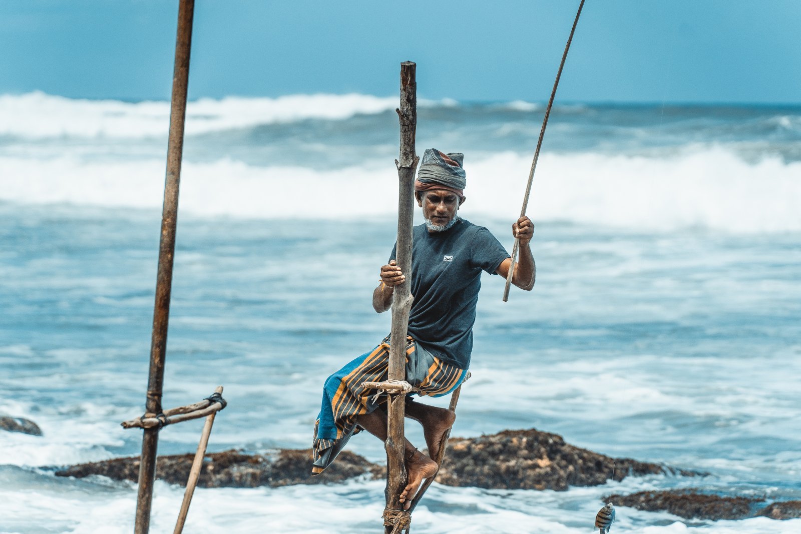 Fisherman standing on a wooden pole in the ocean, holding a fishing rod, with waves crashing behind.